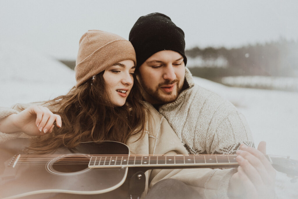 dívka a kluk sedí u ohně na sněhu v zimě a kluk jí učí hrát na kytaru, padá sníh