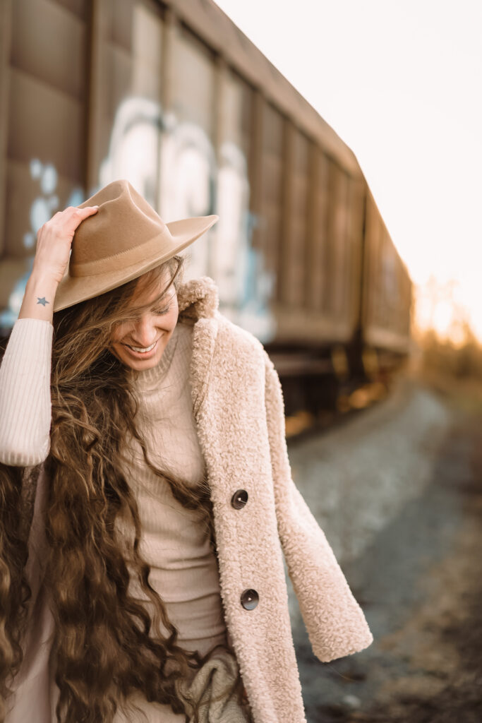 žena s dlouhými vlasy s béžovým kloboukem se směje a za ní projíždí nákladní vlak a za vlakem prosvítá sluníčko